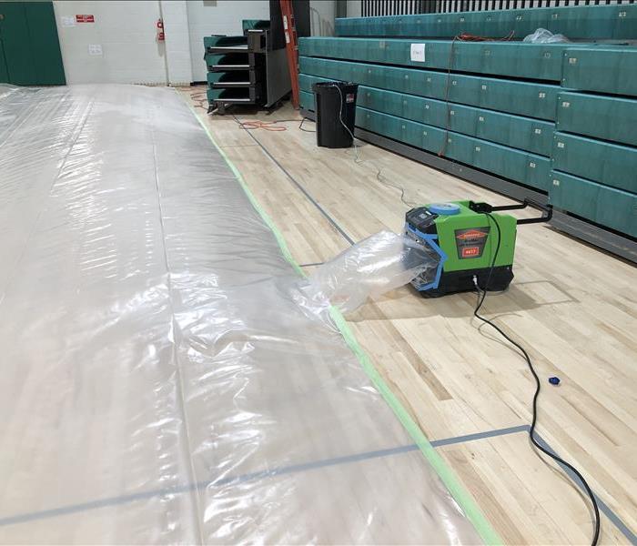 Gymnasium floor with clear plastic tenting and a desiccant machine blowing air under the plastic to dry the floor